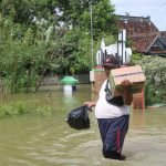 Foto : Sunardi sedang Memikul Bahan Pokok di Tengah akses jalan Desa Karangrowo yang tergenang Banjir. (Dok. Mitrapost.com/Ilham)